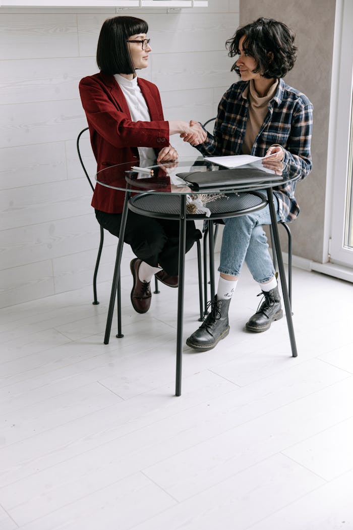 Two women sitting at a glass table indoors, shaking hands over business documents.